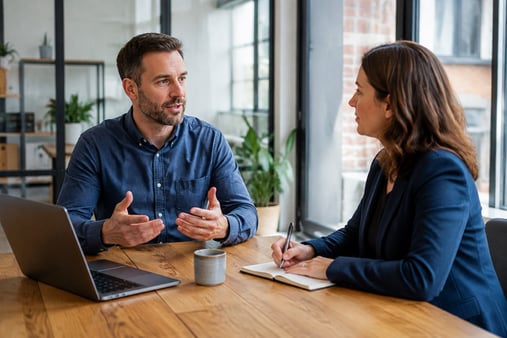 Two people discuss feedback at an office table while one takes notes beside a laptop.