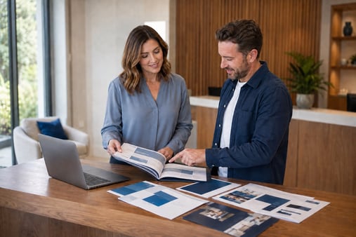 Two people review an open planning folder beside a laptop and printed layouts in a bright reception workspace.
